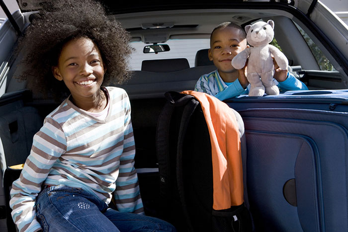 Two kids smiling from back of family SUV with luggage and toy